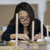 A woman with black hair and glasses works at a table with small pieces of cardboard.