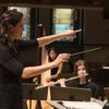 A woman conducts an orchestra with students playing flute and harp in the background.